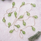 A garland of small, translucent light green glass ornaments hanging from a natural jute string, laid out on a pale textured surface with a pine branch in the corner.