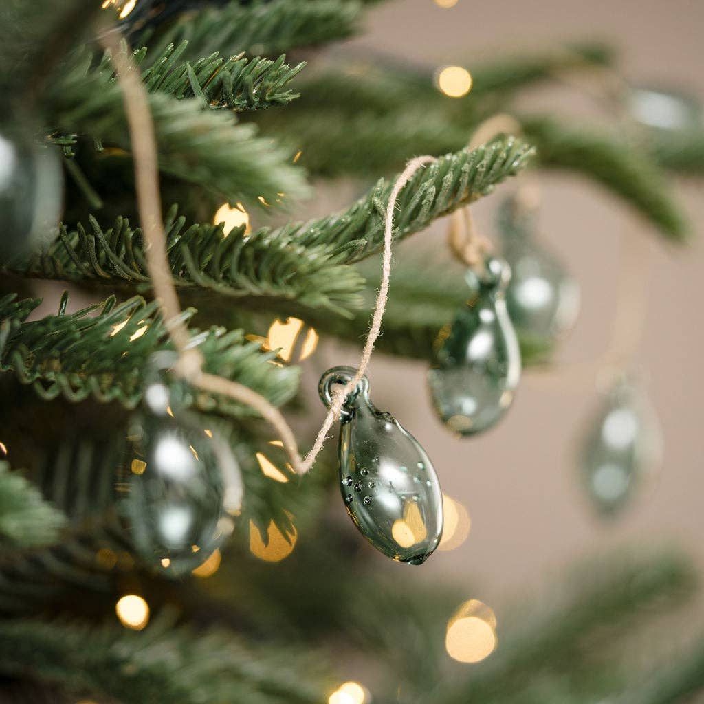 A close-up of a green glass teardrop Christmas ornament hanging from a pine tree branch with twine, alongside warm white bokeh string lights.
