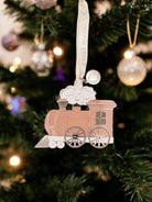 Decorative train ornament hanging on a Christmas tree with lights and ornaments in the background.