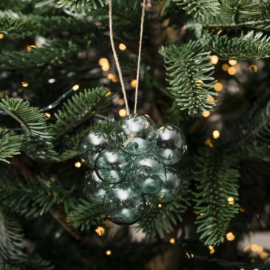A cluster of clear, textured glass Christmas ornaments hanging from a natural twine string on a green pine tree with warm yellow string lights.