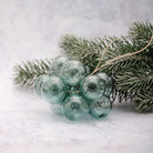 A cluster of translucent dusty blue-green glass ornaments shaped like pine berries, attached to a sprig of artificial evergreen branches on a light gray textured surface.
