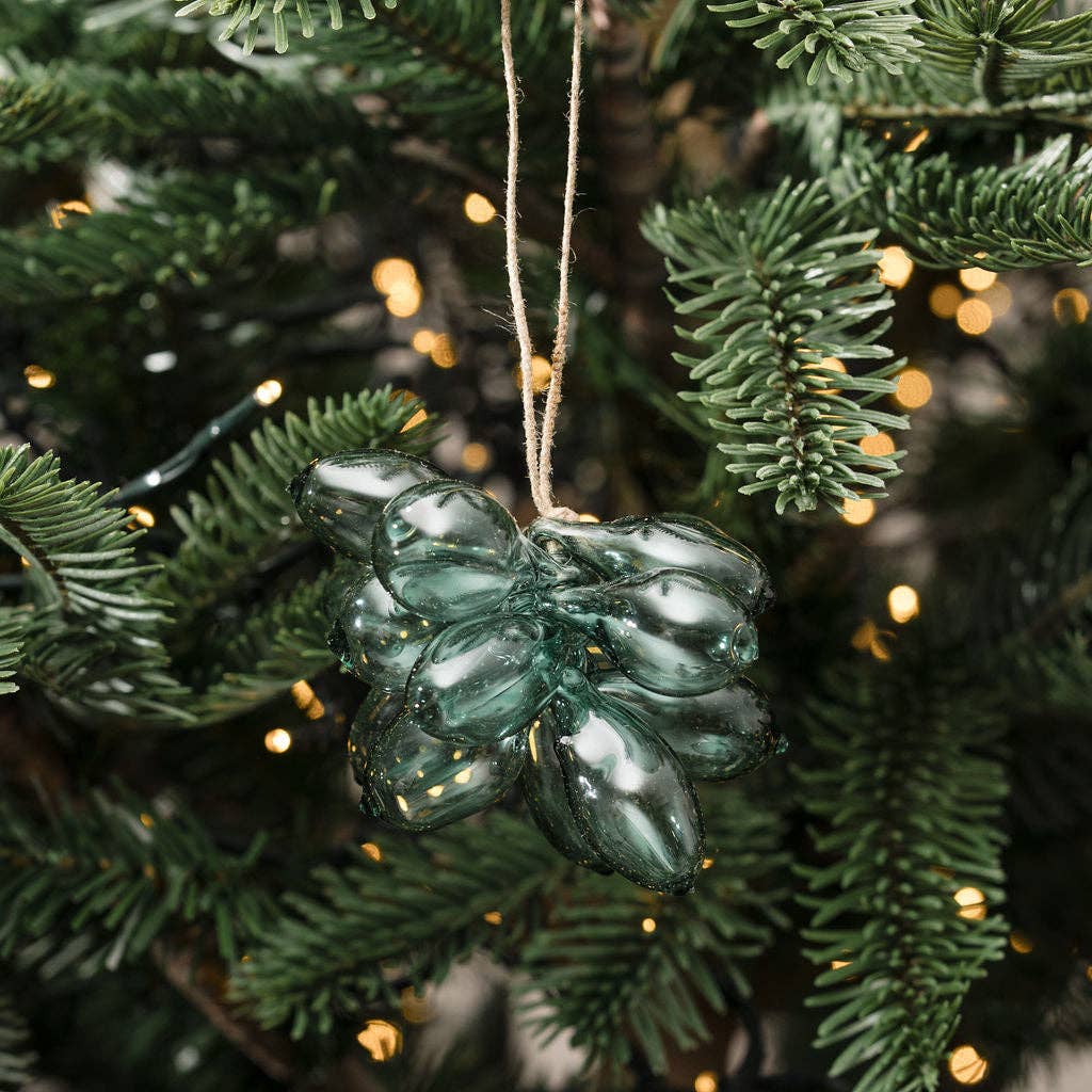 A dark green, bulb-shaped glass Christmas ornament hanging from a jute string on a pine tree with warm bokeh string lights.
