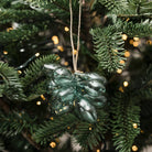 A dark green, bulb-shaped glass Christmas ornament hanging from a jute string on a pine tree with warm bokeh string lights.