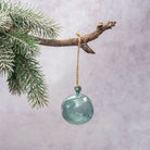 A round, swirled translucent teal glass ornament hanging from a natural twine loop attached to a rustic tree branch with snowy green pine needles, set against a soft textured light gray background.
