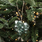 A green glass ornament shaped like a cluster of grapes hangs from a string on a pine tree with warm string lights in the background.