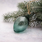 A translucent teal glass ornament hanging from a rustic twine string, nestled among frosted green pine branches against a soft, light gray background.