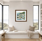 Framed artwork of a coastal scene on a wall above a window seat with pillows and a blanket.